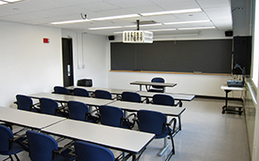 This photo shows the room the lectures were given in with rows of tables and moveable chairs for a maximum of 20 students.  There is an overhead and manual slide projector, a large chalkboard at the front and a window to the students’ right.