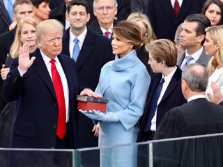 With his right hand raised in the air, an old, reddish-grey haired man rests his left hand on a Bible held by an elegantly-dressed young woman.