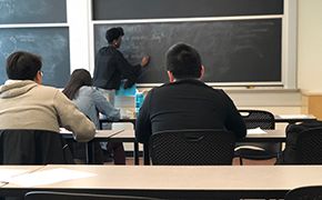 View from back of a recitation classroom. Three students sitting at tables. A teaching assistant writing on chalkboard.