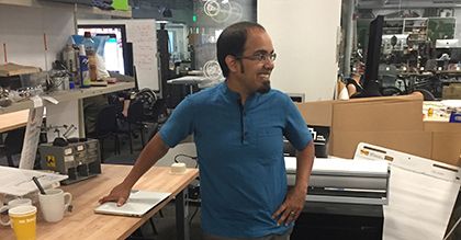 Image of a man standing in front of a wooden and metal work bench. There are monitors and assorted materials in the background.