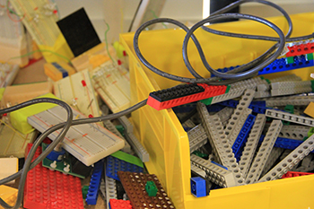 Yellow box filled with grey and blue Legos. Other Legos of various shapes and colors on table outside of box. A grey cable rests on top of the Legos. Grey circuit board visible.