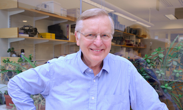 Man in a blue shirt sounding outside a lab. The man has one arm positioned on his hip. He is smiling.