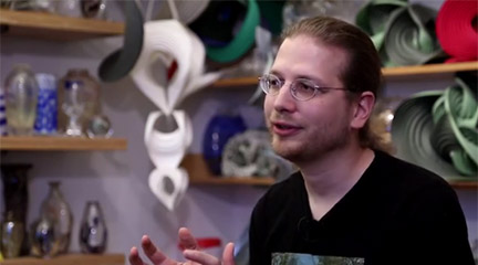 Man with glasses sits in front of geometric paper sculptures.