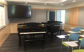 Classroom with two black pianos positioned near a whiteboard marked with musical scores. Tablet armchairs are positioned near the pianos. A flat-screen television is mounted to the wall.