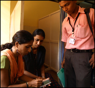 A female health worker logs data on a device as two others look on.